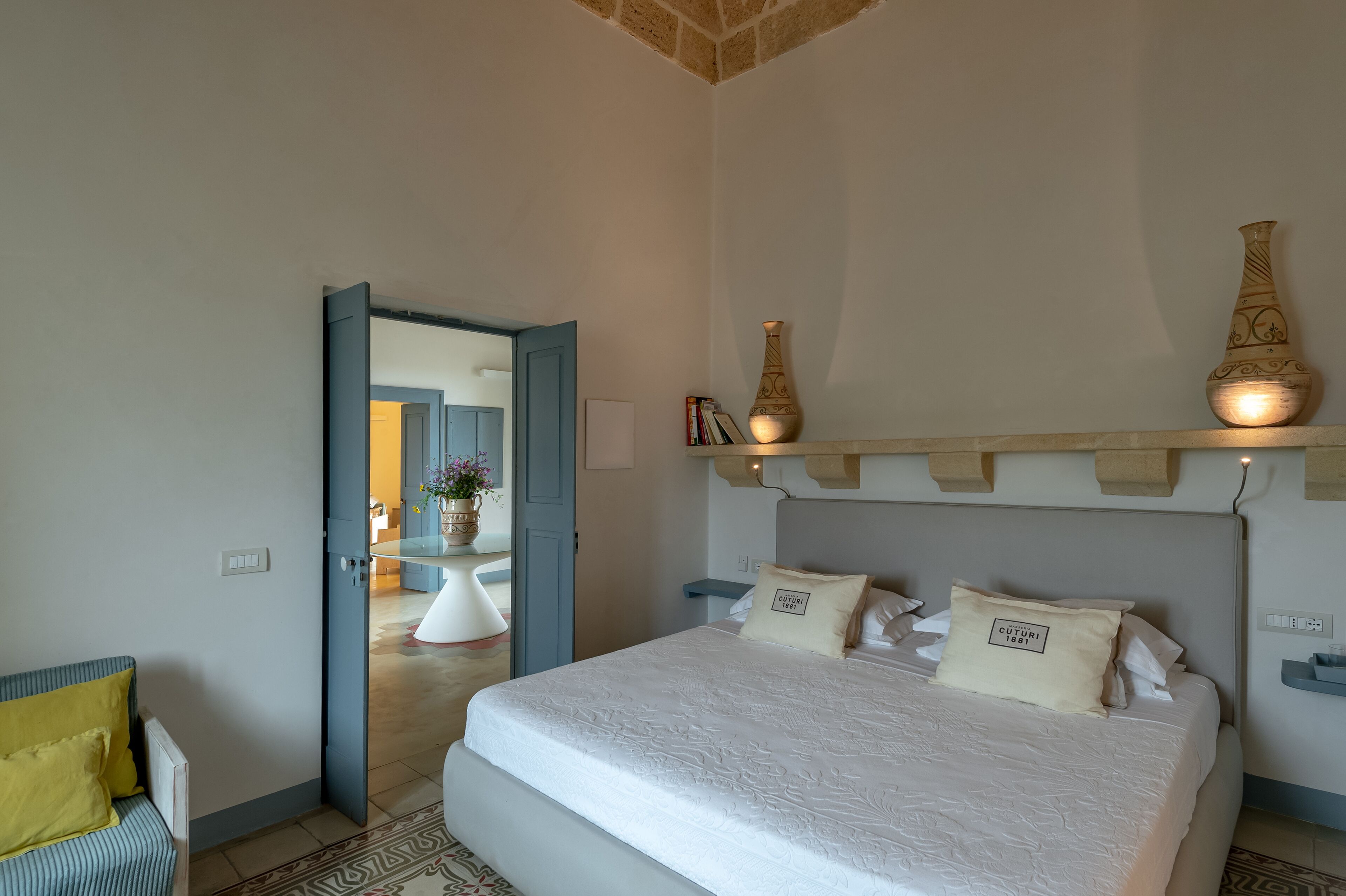 A bedroom with a large bed, a bedside table, a nightstand, and a shelf with decorative items.