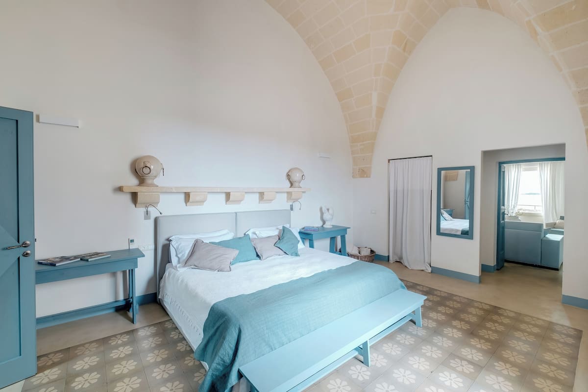 A bedroom with a bed, bedside tables, a bench, a mirror, and a door.