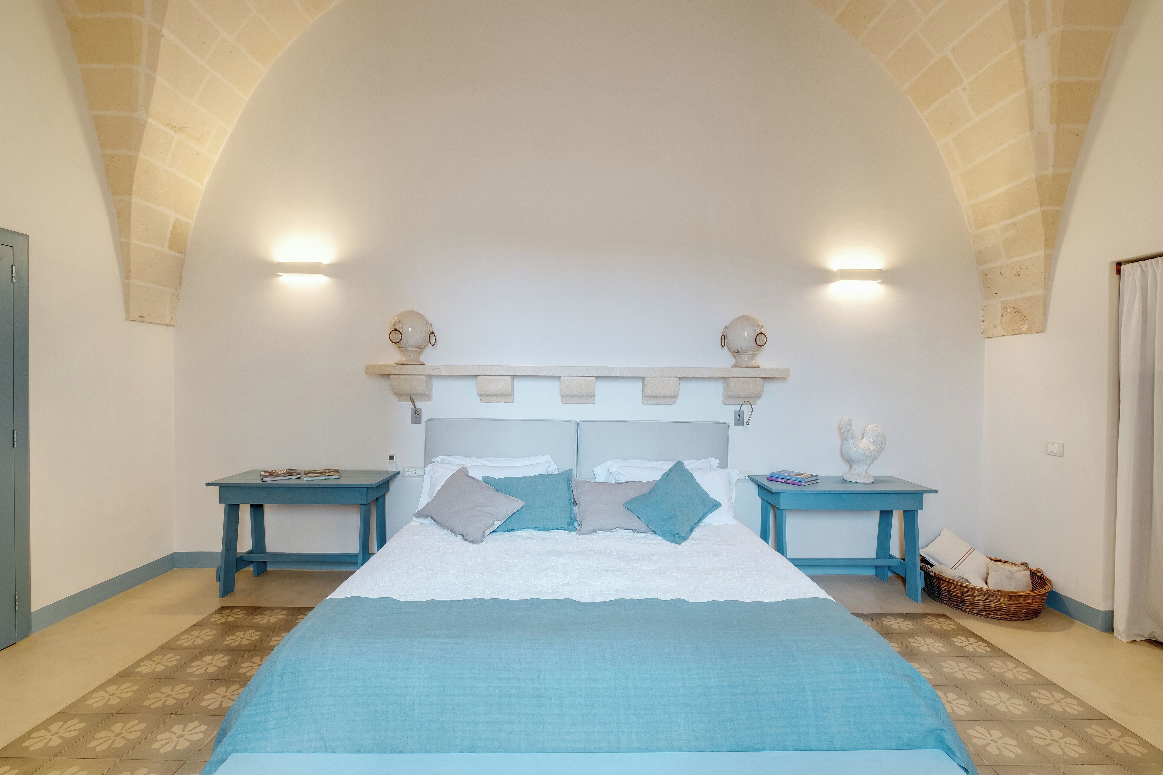 A bedroom with a large bed, two bedside tables, a nightstand, a wall-mounted shelf, and a patterned floor.