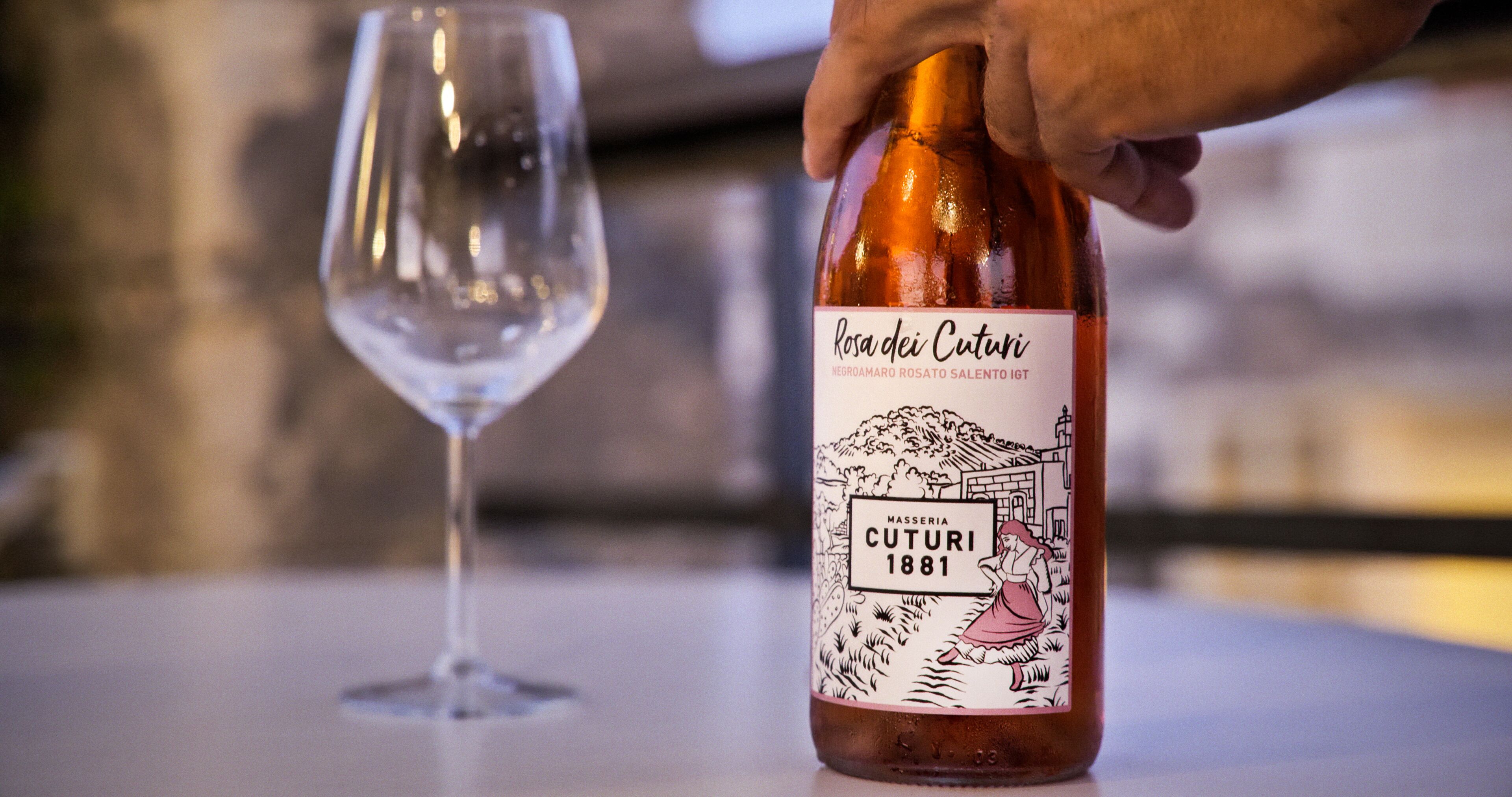 Une bouteille de vin Rosa dei Cuturi, dont l’étiquette représente une femme et un paysage, est posée à côté d’un verre à vin vide sur une table.