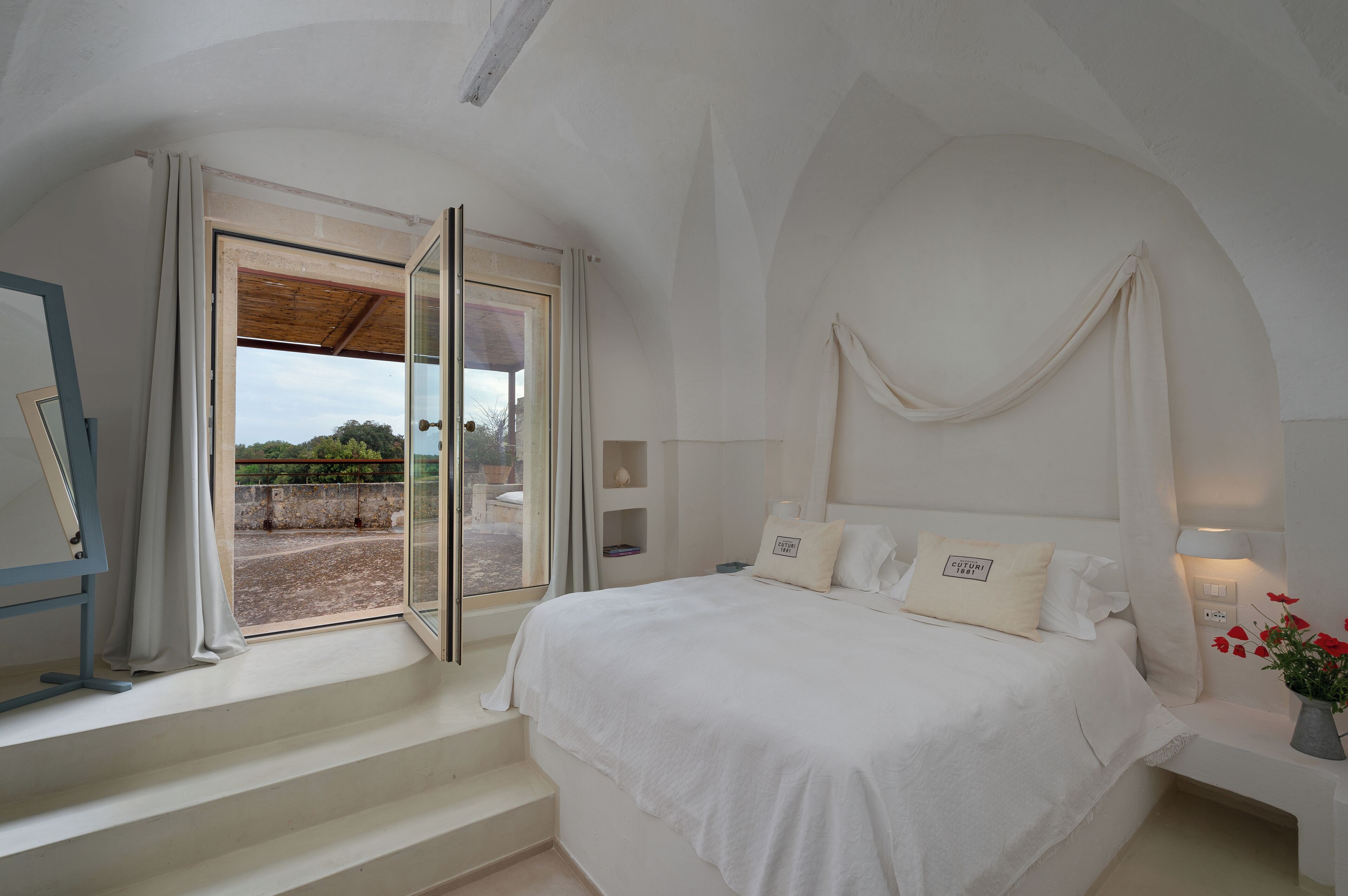 Ein Schlafzimmer mit einem großen Bett, einem Fenster mit Vorhängen, einem Nachttisch mit Lampe und Blick ins Freie.