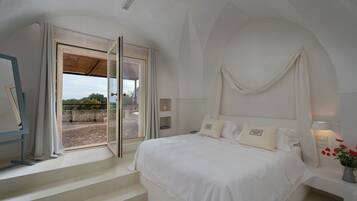 Ein Schlafzimmer mit einem großen Bett, einem Fenster mit Vorhängen, einem Nachttisch mit Lampe und Blick ins Freie.