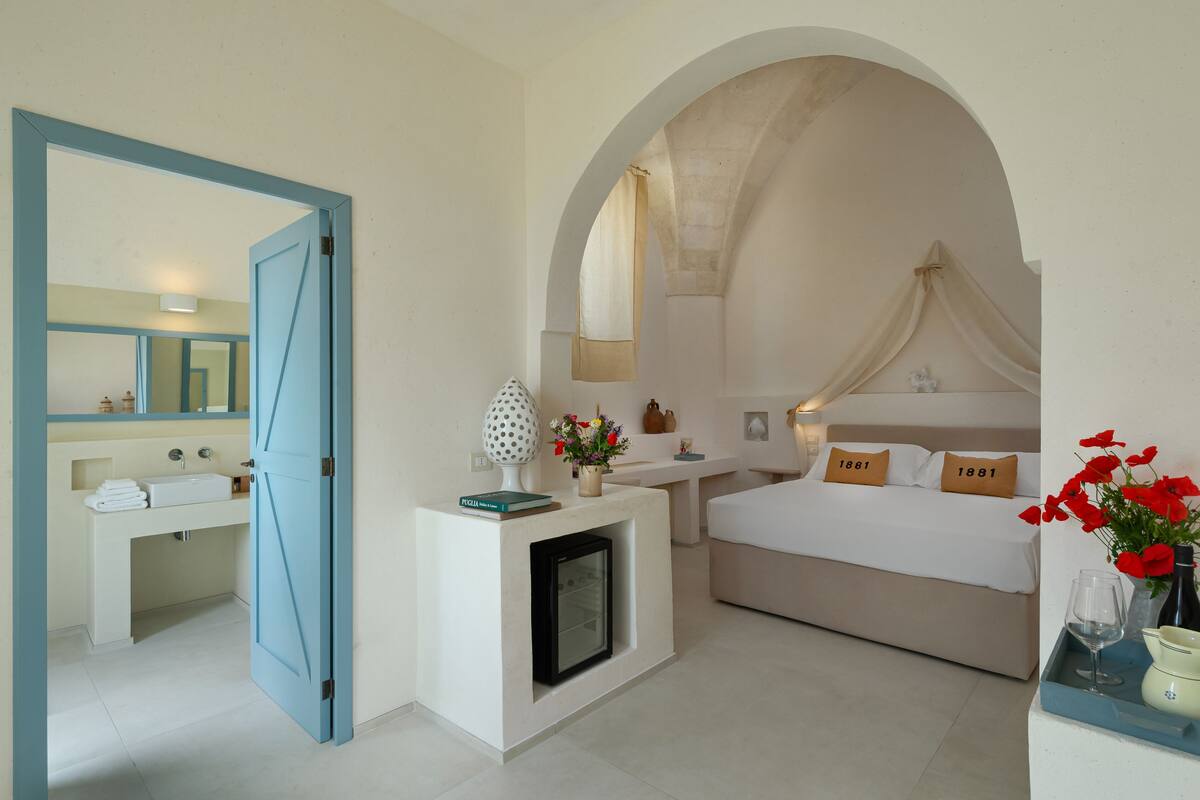 A bedroom with a bed, a bedside table, a nightstand, a mirror, a window with curtains, and a small fireplace.