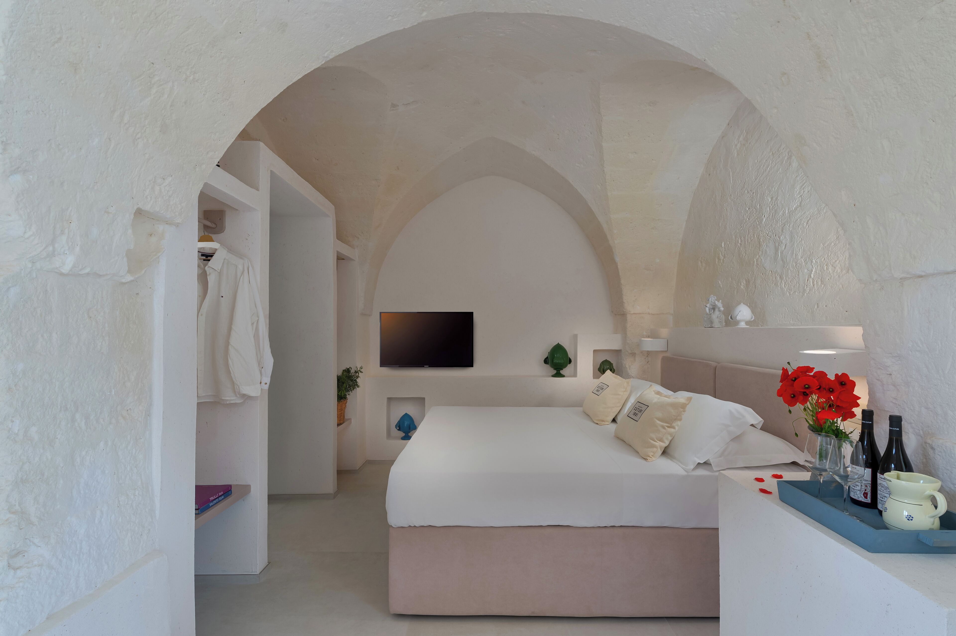 A bedroom with a large arched doorway, a bed with white bedding, a flat-screen TV on the wall, a small plant, and a vase with red flowers on a bedside table.