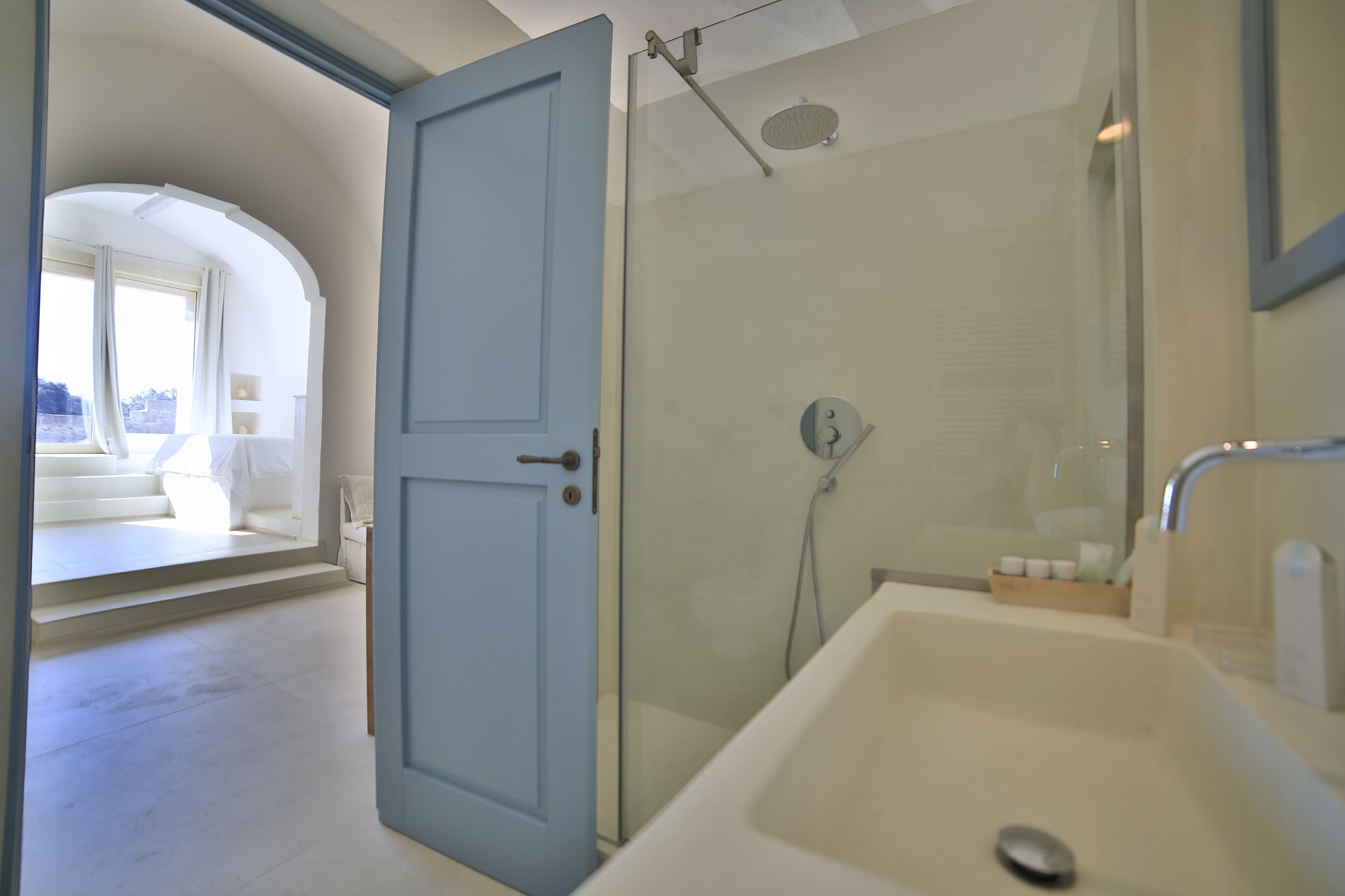 A modern bathroom with a glass-enclosed shower, a white bathtub, and a view of a balcony with a white sofa.