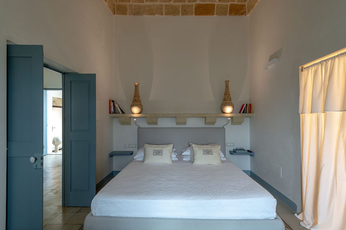 A bedroom with a bed, bedside tables, a shelf with decorative items, and a door leading to another room.