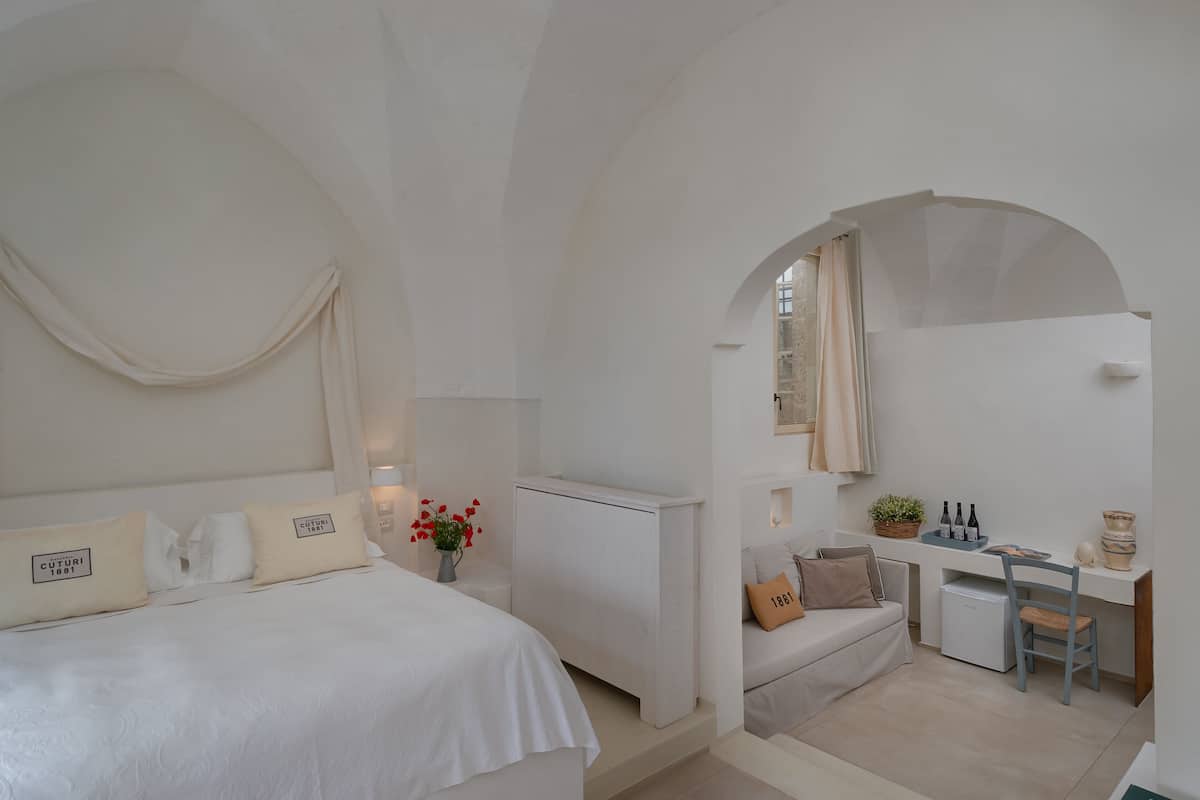 A minimalist bedroom with a bed, bedside table, and a small desk area with a chair and a plant.
