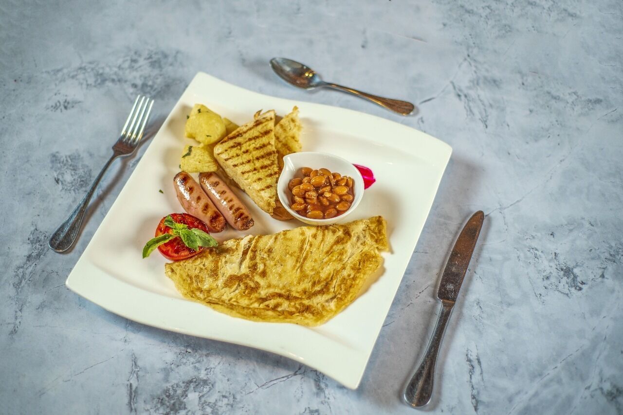 Un plato con salchichas a la parrilla, patatas asadas, un tomate a la plancha, un tazón de frijoles y una tortilla doblada.