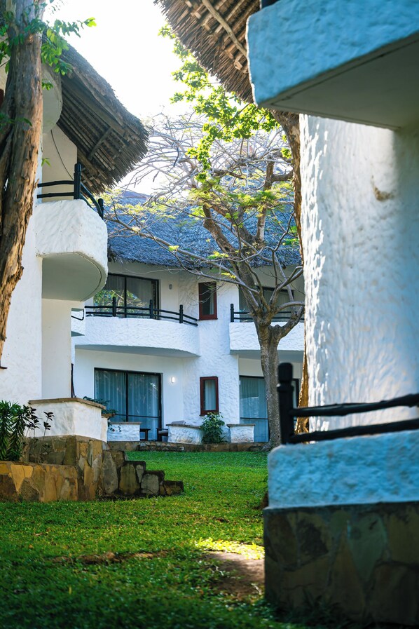 A courtyard with white buildings, a tree, and a stone bench. - Safari Beach Hotel  (Diani Beach)