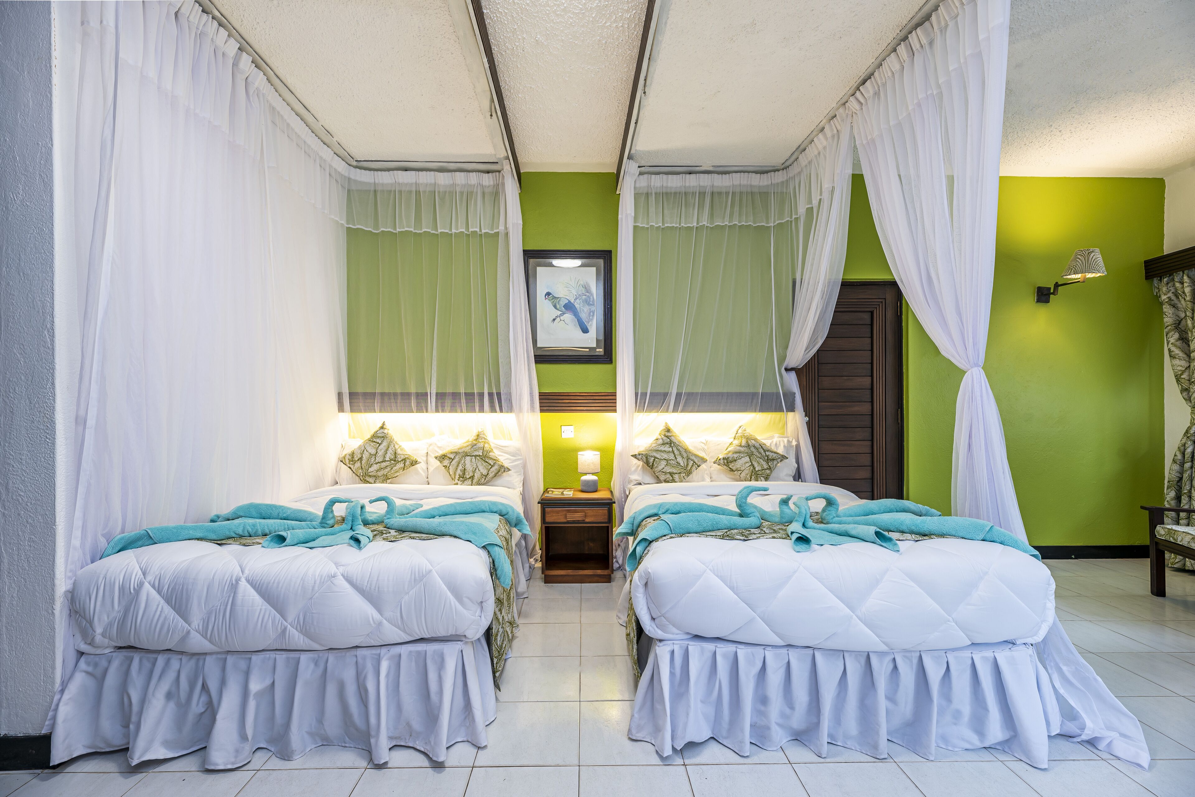 A room with two beds, each with white bedding and teal accents, a nightstand with a lamp, and a framed picture on the wall.