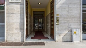 A building entrance with a marble facade, a sign reading "VIA CALIFORNIA", and a red carpet leading to a door.