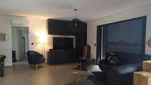 A modern living room with a flat-screen TV, a dark sofa, a patterned coffee table, and a large window with blinds. - Villa Moderne Proche Solenzara (Ventiseri)