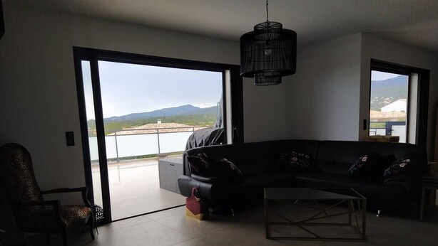 A living room with a sofa, a coffee table, and a chair. There is a large window with a view of a body of water and mountains. - Villa Moderne Proche Solenzara (Ventiseri)
