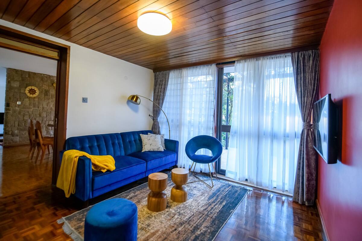 Uma sala de estar com um sofá azul, uma poltrona azul, uma mesa de centro de madeira e uma janela grande com cortinas leves.