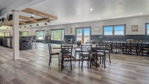 Un vaste espace dédié à la salle à manger, avec des tables et des chaises en bois, de grandes fenêtres offrant une vue dégagée et, au fond, une cuisine moderne.