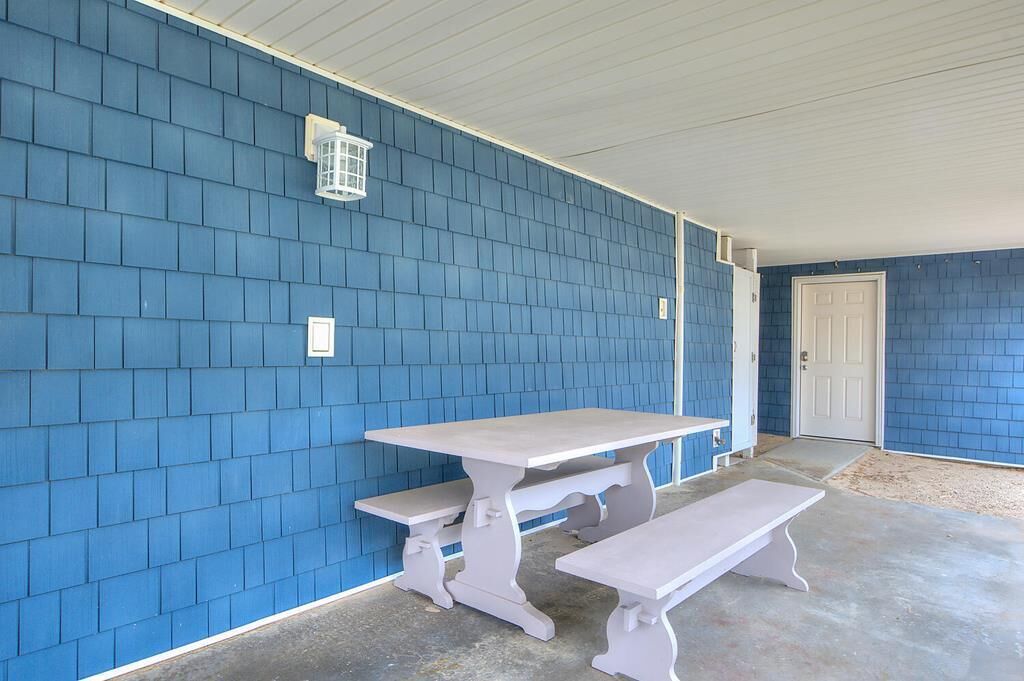 Uma área de varanda coberta com uma mesa de piquenique branca e bancos, paredes com revestimento azul e uma porta branca.