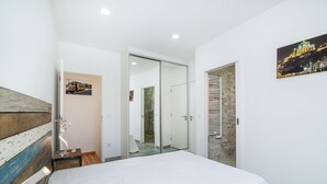 Una camera da letto con un letto, una testiera in legno, una porta vetrata che dà su un bagno e un dipinto che raffigura uno skyline urbano.