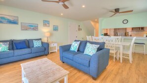 A living room with two sofas, a coffee table, and a dining area with white chairs and a table. There are ceiling fans, a clock, and a kitchen in the background. - Oceanfront Codfather Unit 1, a spacious 3-bedroom duplex in Oak Island. (Oak Island)