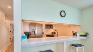 A compact kitchen with a refrigerator, microwave, and coffee maker. There are two stools and a clock on the wall. - Oceanfront Codfather Unit 1, a spacious 3-bedroom duplex in Oak Island. (Oak Island)