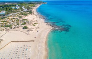 Vue aérienne d’une plage aux eaux turquoise et limpides, bordée d’un rivage sablonneux et ponctuée de quelques parasols.