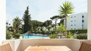 A wooden table and chairs on a balcony overlooking a swimming pool with lounge chairs and umbrellas, surrounded by greenery and buildings. - Alvaflor 102 - Vilamoura by HD Properties (Vilamoura)