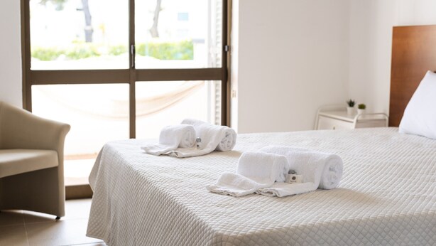 A hotel room with a bed, a chair, a nightstand, and a window with a view of greenery. - Alvaflor 102 - Vilamoura by HD Properties (Vilamoura)