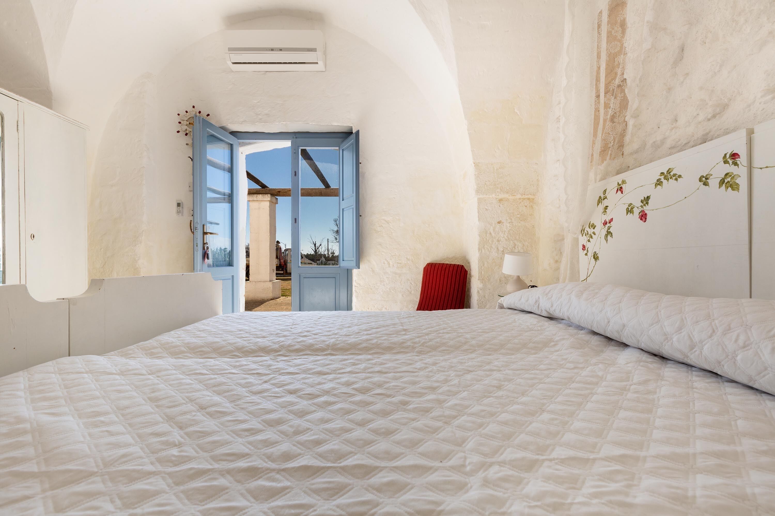 A bedroom with a large bed, a red chair, a white wall with a floral design, and an open door leading to a balcony.