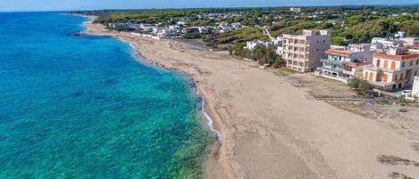 A coastal area with a sandy beach, clear blue waters, and buildings with balconies overlooking the sea.