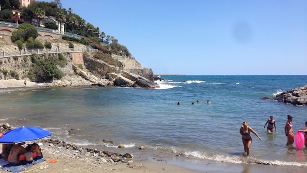 Une plage au sol rocheux, un ciel dégagé et quelques personnes dans l’eau.