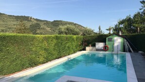 Une piscine à l’eau d’un bleu limpide, entourée d’une terrasse en pierre. On y trouve un petit abri surmonté d’un drapeau, et une haie en arrière-plan.