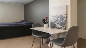 A modern room with a bed, a desk, two chairs, a small potted plant, and a mountain landscape painting. - Studio-sleeps 4-city Centre-parking-evcharger (Tromsø)