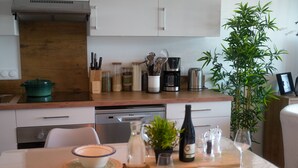 Una cocina moderna con encimera de madera, cafetera, tostadora y lavavajillas. Hay una mesa de comedor preparada con un tazón, botella de vino, copas y cubiertos.