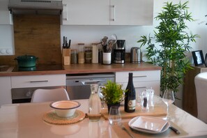 A modern kitchen with a wooden countertop, a coffee maker, a toaster, and a dishwasher. There is a dining table set with a bowl, wine bottle, glasses, and cutlery.