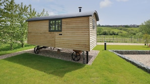 A wooden carriage-like structure with a window and a chimney, situated on a grassy area with a stone wall and a fence in the background. - Malbec (Ryde)
