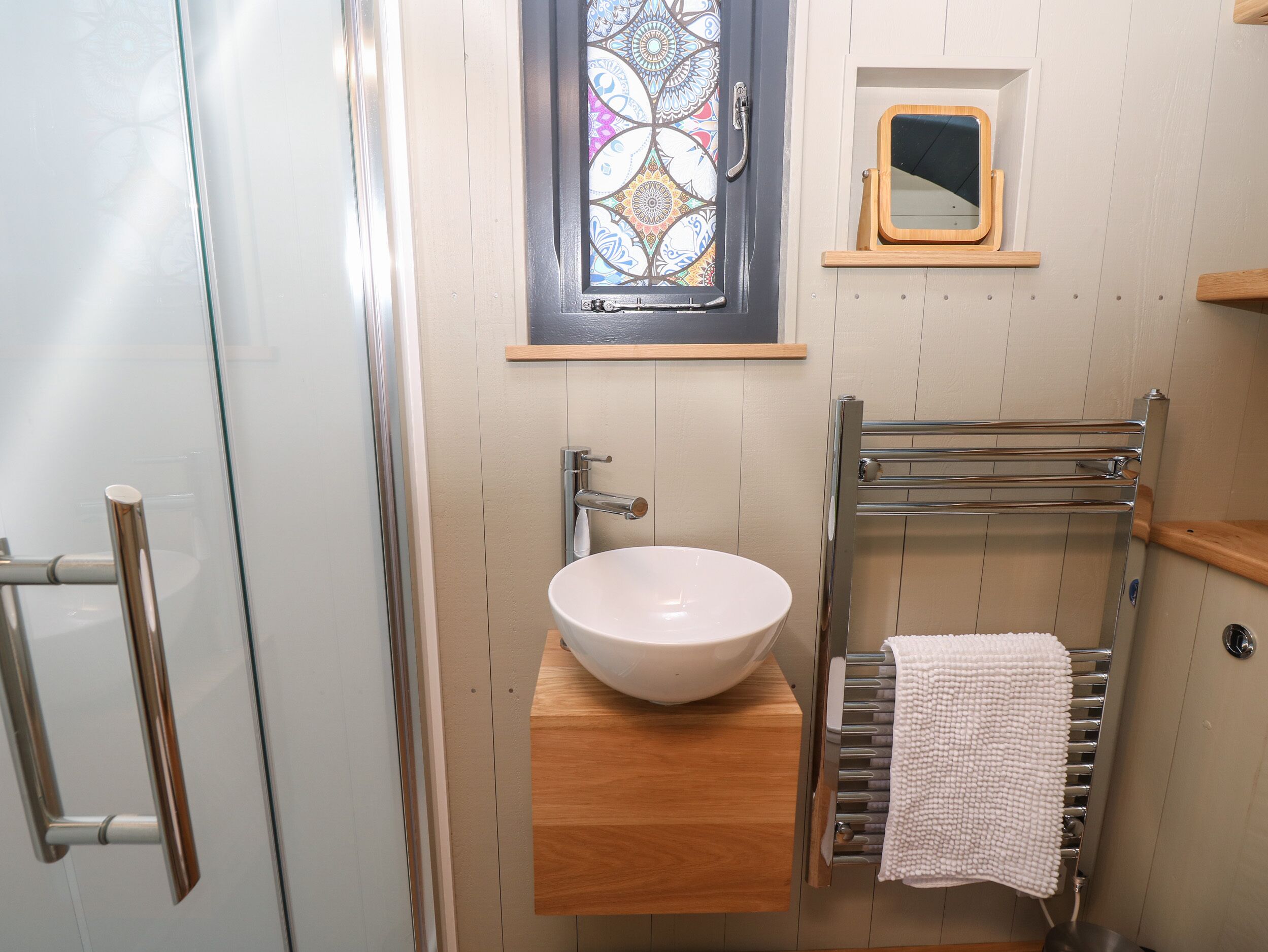 A bathroom with a glass shower enclosure, a white sink with a wooden countertop, a towel rack, and a small mirror.