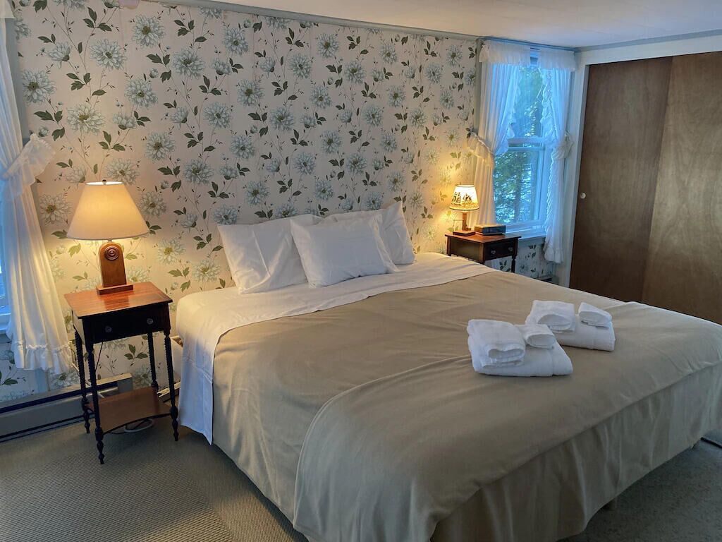 Ein ordentlich bezogenes Bett mit weißen Leinentüchern, zwei Nachttische mit Lampen und einem floralen Wandpapier.