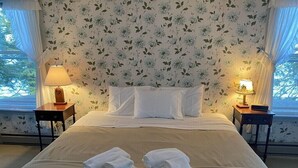 Una cama bien hecha con sábanas blancas y dos toallas dobladas. A cada lado de la cama hay dos mesitas de noche con lámparas. La habitación tiene papel tapiz con motivos florales y una ventana con cortinas.