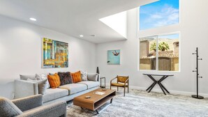 A modern living room with a grey sofa, a coffee table, and a chair. There is a painting on the wall and a window with a view of palm trees.