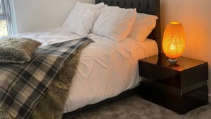 A neatly made bed with white linens and a fluffy blanket, a dark headboard, and a bedside table with a lit lamp. - Immaculate 2-bed Apartment in Grays (Grays)