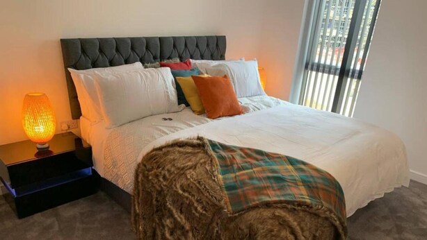 A bedroom with a tufted headboard, a bed with white linens, colorful pillows, a plaid throw blanket, and a nightstand with a lamp. - Immaculate 2-bed Apartment in Grays (Grays)