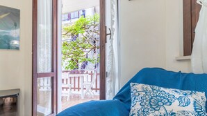 A room with a blue sofa, a patterned pillow, a glass door leading to an outdoor balcony with chairs and a tree. - Italian Experience - Conero Garden Apt (Marcelli)