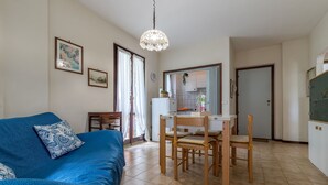 A cozy living room with a blue sofa, a dining table with chairs, a kitchen area, and a large window with curtains. - Italian Experience - Conero Garden Apt (Marcelli)