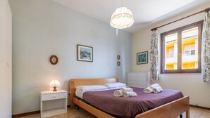 A bedroom with a wooden bed, a bedside table with a lamp, a window with curtains, and a view of a yellow building outside. - Italian Experience - Conero Garden Apt (Marcelli)