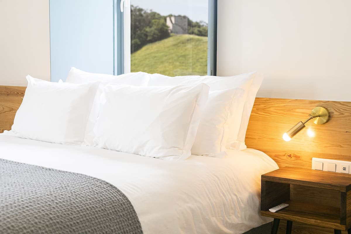 Una cama bien hecha con sábanas blancas, cabecera de madera y mesita de noche con lámpara.
