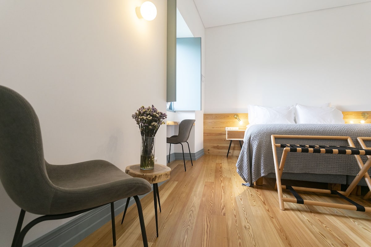 Una habitación de hotel moderna con suelo de madera, una silla tapizada de color gris, una pequeña mesa redonda con un jarrón, una cama con colcha a rayas grises y blancas, y un banco de madera con cojín a rayas negras y blancas.