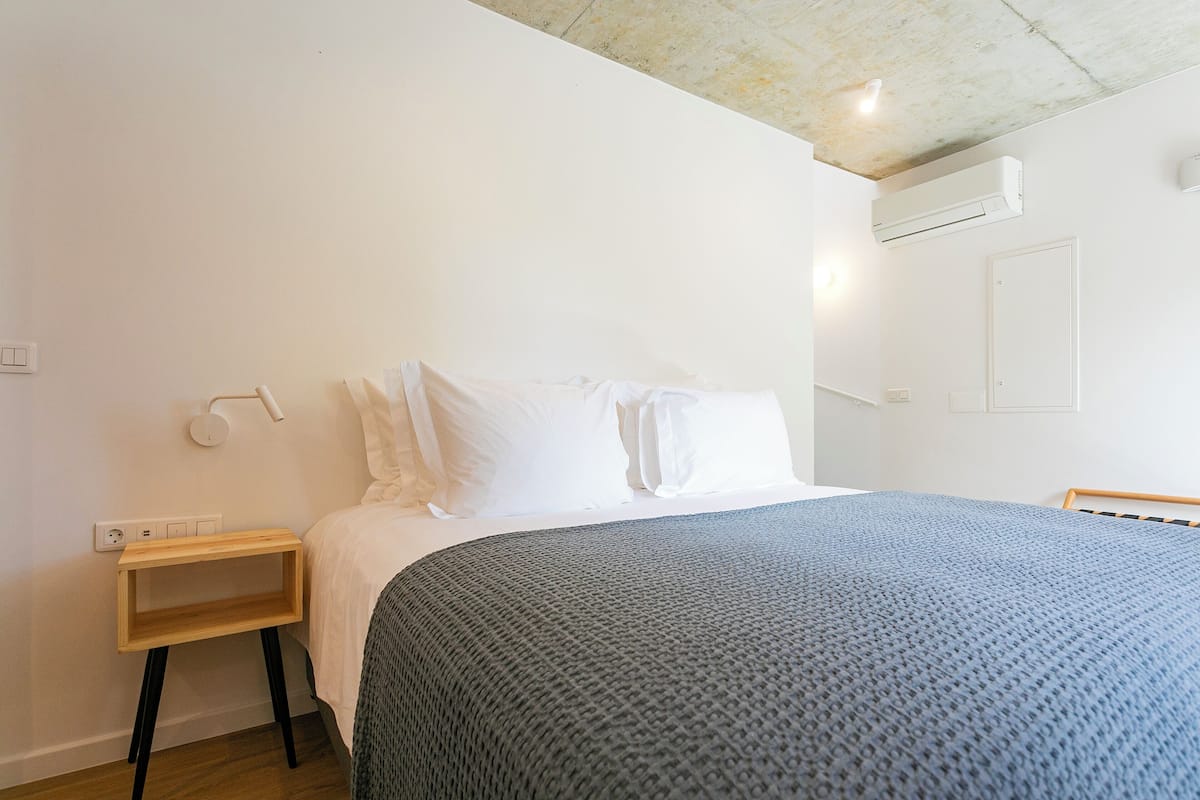 Un dormitorio con cama, almohadas blancas, manta gris, mesita de noche de madera y pared blanca con unidad de aire acondicionado.