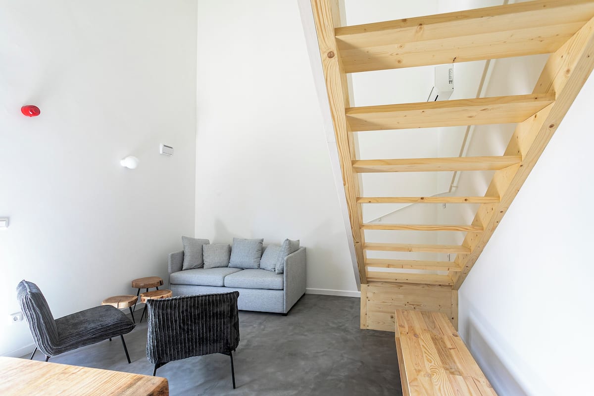 Una sala de estar moderna con un sofá gris, dos sillones, una mesa de madera y una escalera de madera.