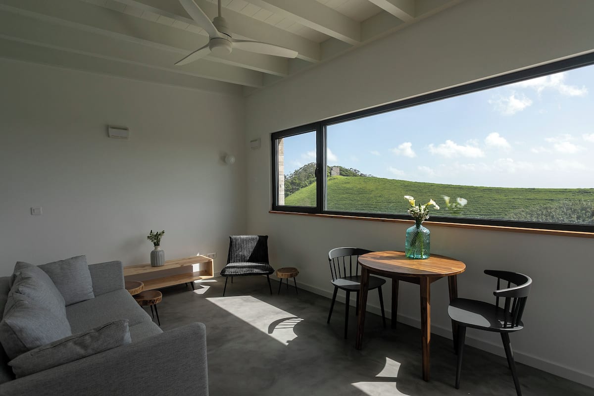 Una sala de estar moderna con un ventanal grande, un sofá gris, una mesa de madera y un florero con flores.