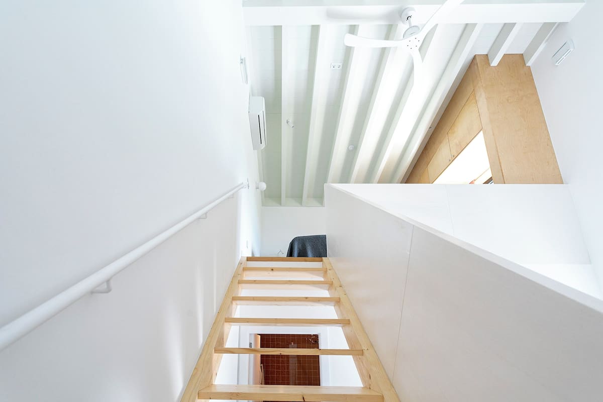 Una escalera estrecha con vigas de madera, paredes blancas y una habitación con tejas rojas visible abajo.
