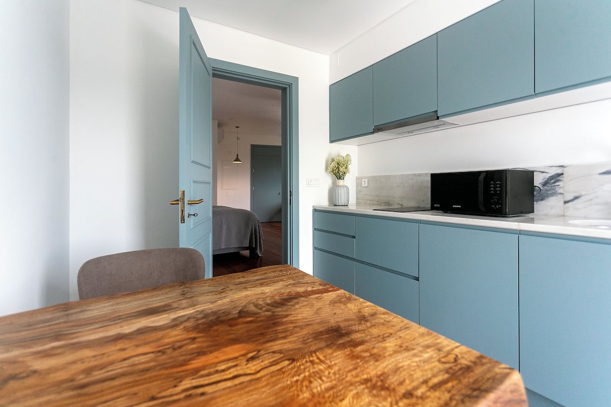 Una cocina compacta con armarios verde azulado, una mesa de comedor de madera y un microondas en la encimera.